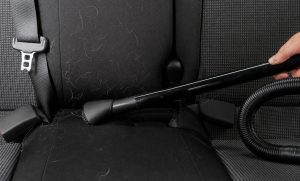 Removing light-colored pet hair from a black cloth car seat with a vacuum brush tool, car interior detailing and cleaning.